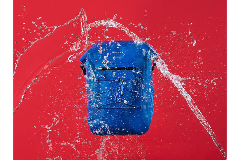 A blue Spinlock waterproof bag being splashed with water against a red background photographed by Richard Boll.